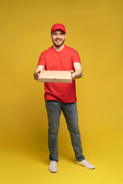 Happy man from delivery service in red t-shirt and cap giving food order and holding pizza box isolated over yellow - Fotoğraf, Görsel