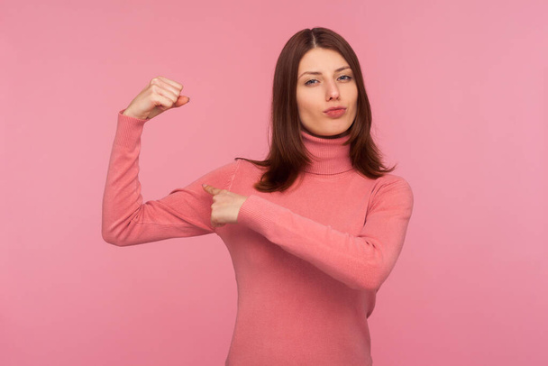 Silna, pewna siebie brunetka w różowym swetrze, pokazująca mięśnie rąk podnoszące rękę, wolną i niezależną, emancypację. Kryty studio strzał izolowany na różowym tle - Zdjęcie, obraz
