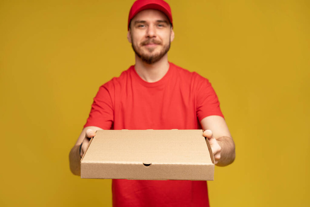 Happy man from delivery service in red t-shirt and cap giving food order and holding pizza box isolated over yellow - Photo, image