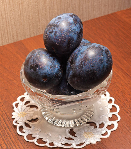 Ciruelas azules en un vaso de vidrio.El jarrón está sobre una servilleta en la mesa.Los frutos están maduros y jugosos.. - Foto, Imagen