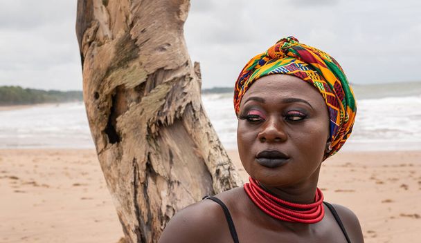Ghanaian Αφρικανική γυναίκα με πολύχρωμο φόρεμα κεφαλής κάθεται σε ένα σπασμένο κούτσουρο φοίνικα στην παραλία στο Axim Γκάνα Δυτική Αφρική - Φωτογραφία, εικόνα