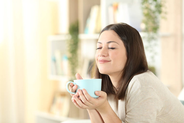 Задоволена жінка розслабляється тримає чашку кави, сидячи на дивані у вітальні вдома
 - Фото, зображення