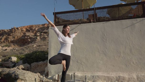 Молода жінка практикує йогу в сонячний день на скелястому узбережжі моря.
 - Фото, зображення
