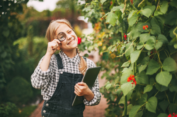 Egy gyönyörű tinédzser lány táblagéppel a kezében megvizsgál egy növénymintát egy nagyítón keresztül. Különös gyerekek.. - Fotó, kép