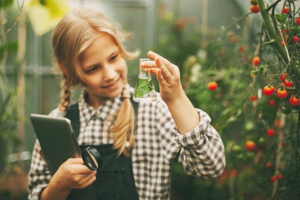 Une belle petite adolescente avec une tablette dans les mains examine un échantillon d'une plante à travers une loupe. Des enfants curieux. Produits biologiques purs - Photo, image