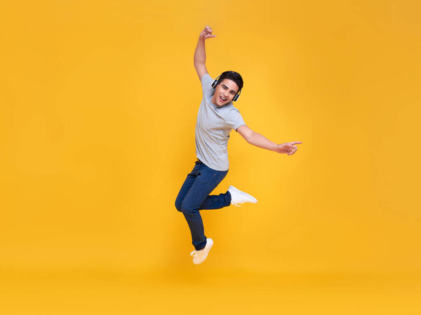 Young handsome Asian man smiling and jumping wearing wireless headphone listening to music isolated over yellow background. - Valokuva, kuva