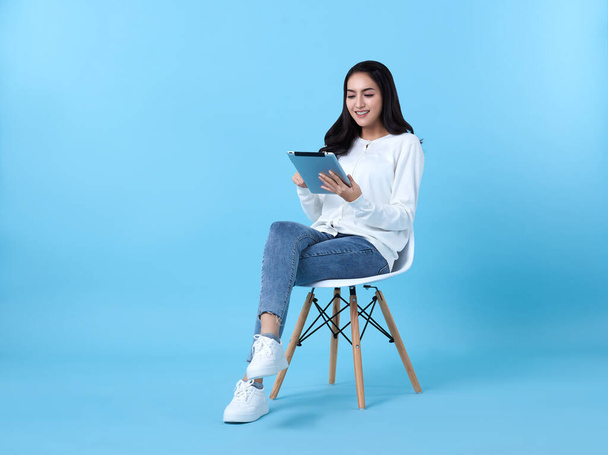 Young woman asian happy smiling in casual white cardigan with denim jeans.While her using tablet computer sitting on white chair isolate on bright blue background. - Fotó, kép