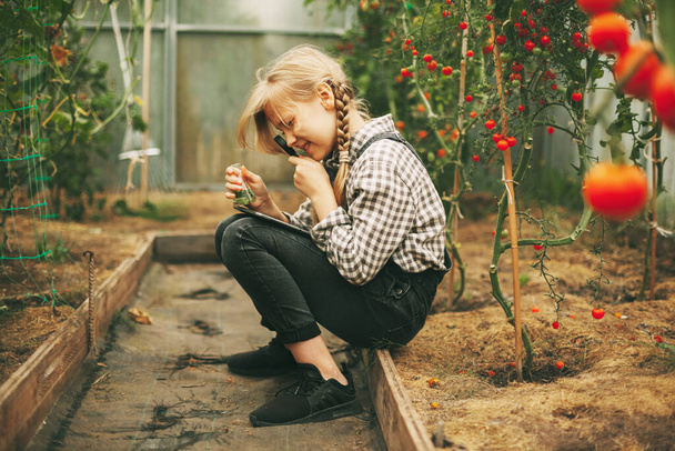 Красива маленька дівчинка-підліток з таблеткою в руках досліджує зразок рослини через збільшувальний келих. Цікаві діти
. - Фото, зображення
