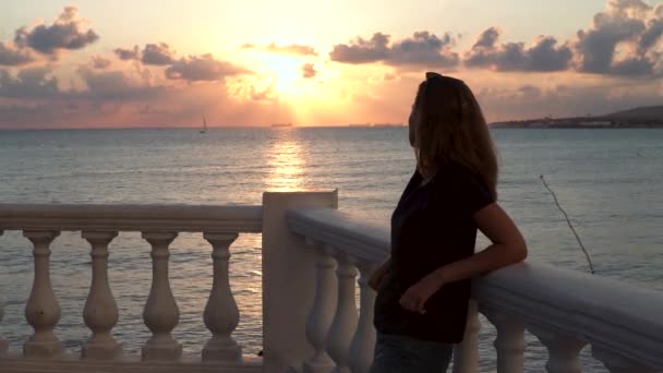 Mujer de pie en el terraplén junto a la orilla del mar y mirando la impresionante puesta de sol. Medios. Chica romántica joven y solitaria apoyada en la barandilla.. - Imágenes, Vídeo