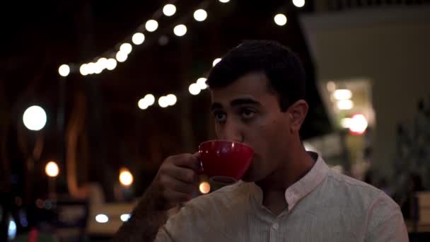悲しい独身ロマンチックな男は、屋外のレストランでガールフレンドを待っています。メディア。夜遅くに一人で座っている白いシャツの退屈男の肖像画と赤いカップから熱い飲み物を飲む. - 映像、動画