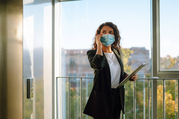 Young business woman  in protective sterile mask talking on mobile phone while standing near modern elevator. Work during pandemic in quarantine city. Covid-19. - Photo, Image