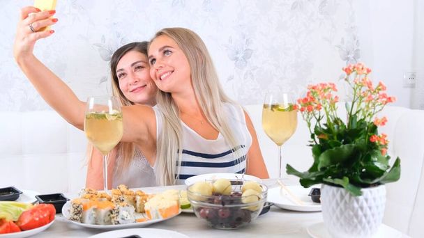  conceito de amizade e festa - duas jovens belas namoradas bebem vinho e tomam selfie no smartphone em uma cozinha acolhedora à mesa, foco seletivo - Foto, Imagem