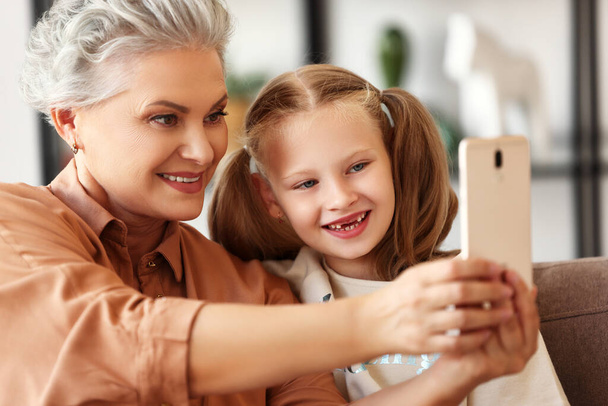 Onnellinen perhe: isoäiti hymyilee ja ottaa selfie iloinen tyttö tyttärentytär istuessaan sohvalla viihtyisässä olohuoneessa hom - Valokuva, kuva