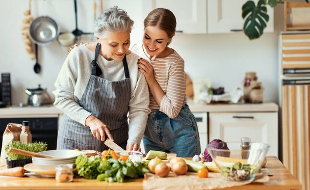 Mutlu aile, neşeli genç kadın olgun anneyi kucaklarken evde taze sebzelerle sağlıklı yemekler hazırlıyor. - Fotoğraf, Görsel