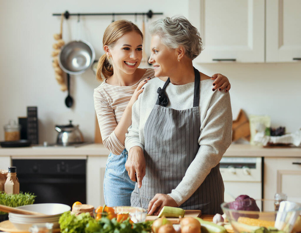 Mutlu aile, neşeli genç kadın olgun anneyi kucaklarken evde taze sebzelerle sağlıklı yemekler hazırlıyor. - Fotoğraf, Görsel