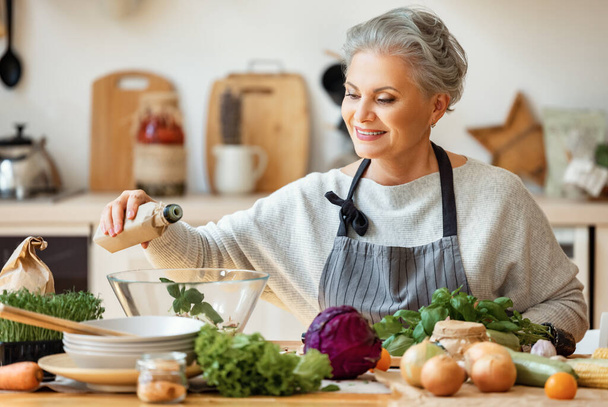 Χαρούμενη μεσήλικη νοικοκυρά με ποδιά χύνει φυτικό λάδι σε μια φρέσκια σαλάτα και κοιτάζοντας την κάμερα ενώ ετοιμάζει υγιεινό πιάτο στην κουζίνα του σπιτιού - Φωτογραφία, εικόνα
