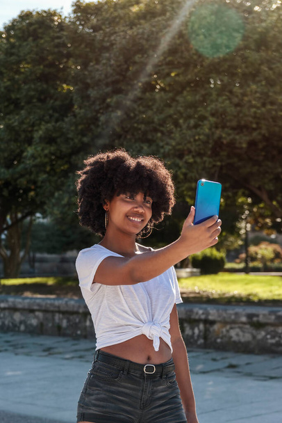 African American girl with afro hair smiles and makes a selfie with her cell phone under the sun walking in an outdoor park - Photo, Image