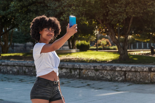 アフリカ系アメリカ人の女の子とともにアフロの髪作りますセルフィーとともに彼女の携帯電話下に太陽の下で歩く屋外公園 - 写真・画像