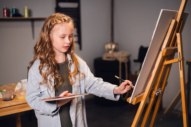 Νεαρή κοπέλα δημιουργικός καλλιτέχνης σε ένα εργαστήριο. Με πάθος ζωγραφίζει μια εικόνα βυθισμένη στη δουλειά. - Φωτογραφία, εικόνα