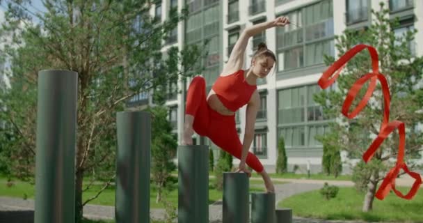 Mladá žena v červeném provádí kallisthenics s stuhou v libing apartmánu dvoře, gymnastka dělá akrobatické cvičení v obytné oblasti, fitness v městském prostředí, 4k 120p Prores HQ - Záběry, video