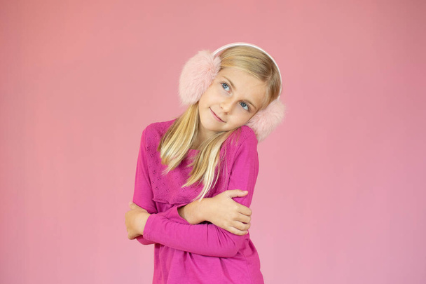 A szőke hajú kislány portréja, boldog mosoly, kék szem, rózsaszín szőrös fülvédőt visel, rózsaszín háttér felett a kamerába néz. - Fotó, kép