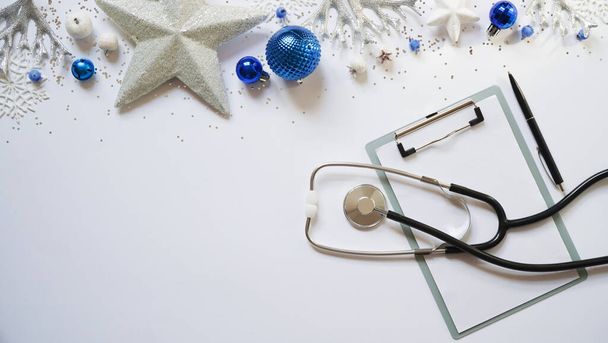 Χριστουγεννιάτικο ιατρικό πανό χριστουγεννιάτικη διακόσμηση λευκά αστέρια, μπλε μπάλες, νιφάδες χιονιού, στηθοσκόπιο και tablet σε λευκό φόντο πάνω άποψη. Αντιγραφή χώρου. Την ιατρική την Πρωτοχρονιά. Ο γιατρός γράφει το κείμενο.. - Φωτογραφία, εικόνα