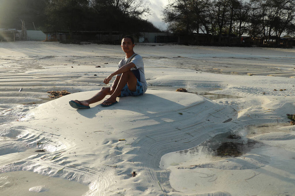 Tinédzser ül fehér homok Tanjung Aan strand, Lombok, Indonézia. Egy fiatal ázsiai férfi nézi a tájat. - Fotó, kép