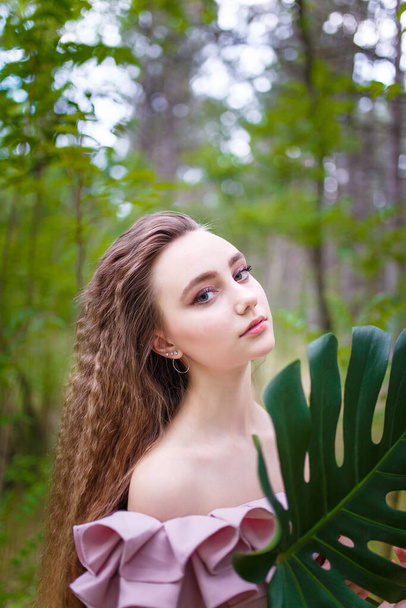 Teenager girl portrait with curly long hair in pink costume among green trees and bushes holding green monstera leaf - 写真・画像