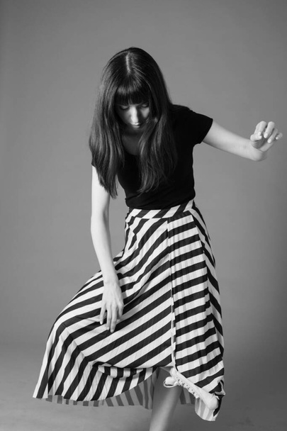 Studio portrait of a pretty brunette woman in a black t-shirt and striped skirt, standing on one foot, against a plain grey background, looking down - Foto, Bild