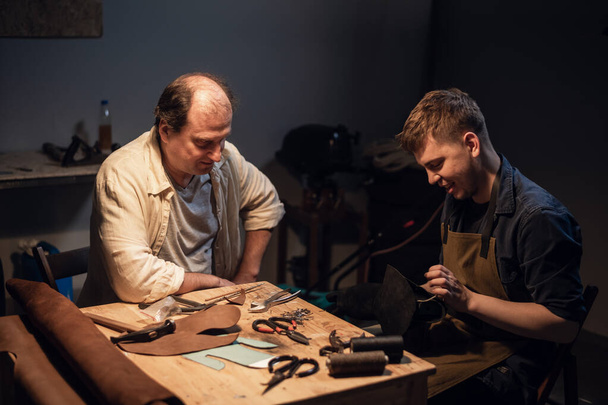 a cute young guy wants to make a gift with his own hands, an experienced shoemaker helps him make shoes out of leather. - Photo, Image