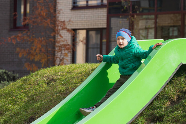 子供たちは屋外遊び場で遊んでいます。緑のコートでアクティブな子供と石のスライドやスイングの帽子。子供のための健康的な夏の活動。小さな男の子が屋外に登る. - 写真・画像