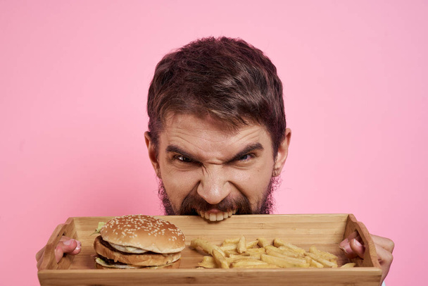 Agressieve kerel met een dienblad in zijn handen hamburger frietjes roze achtergrond hongerige blik - Foto, afbeelding