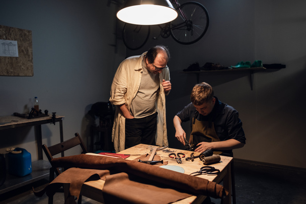 un zapatero anciano y su aprendiz crean zapatos a mano en su taller con diversas herramientas. - Foto, Imagen