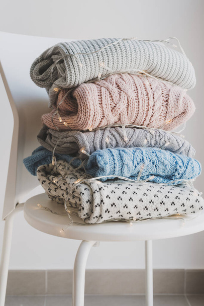 Pile of handmade woolen sweaters on white chair and decorative lights. Warm clothing for cold autumn and winter season - Zdjęcie, obraz