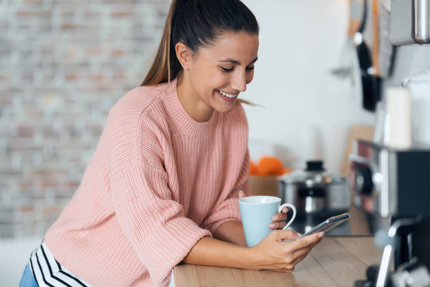 Постріл усміхненої молодої жінки, використовуючи свій мобільний телефон, п'ючи чашку кави на кухні вдома
. - Фото, зображення