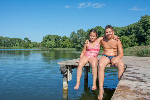 W słoneczny dzień lata, brat i siostra siedzą na moście w strojach kąpielowych na rzece.. - Zdjęcie, obraz