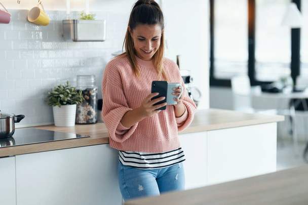 Shot of smiling young woman using her mobile phone while drinking a cup of coffee in the kitchen at home. - Photo, image