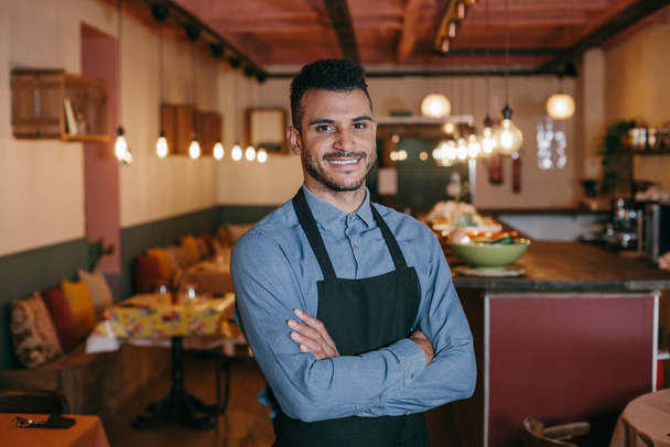 Cheerful positive handsome restaurant owner with crossed arms proud by himself looking at camera. Portrait of an african american attractive waiter or owner, at a restaurant. Small business concept. - Photo, Image