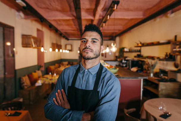 Afrikkalainen amerikkalainen liikemies seisomassa ravintolansa ruokasalissa. Ylpeä ravintolan omistaja kädet ristissä ja katsoo kameraa uhmakkaalla katseella. Pienyrityksen käsite - Valokuva, kuva