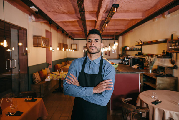 Portrait of young african american businessman standing in the dining room of his restaurant. Proud restaurant owner with his arms crossed and looking at camera. Small business concept - Photo, Image