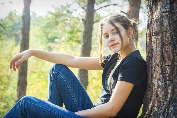 Latem w słoneczny dzień w lesie, dziewczyna siedzi w pobliżu drzewa na trawie. - Zdjęcie, obraz