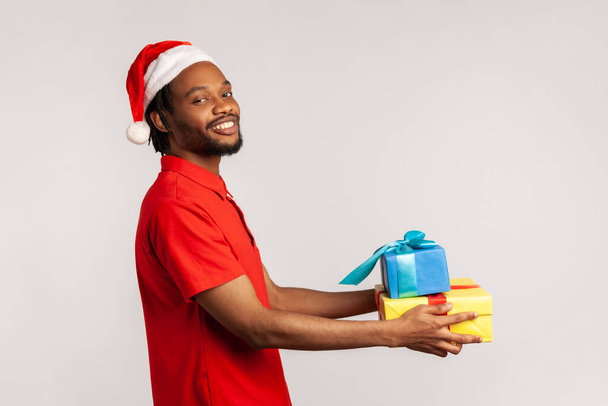 Profile portrait optimistic happy afro-american man with dreadlocks in santa claus hat holding gift boxes looking at camera with toothy smile. Indoor studio shot isolated on gray background - Foto, Imagen