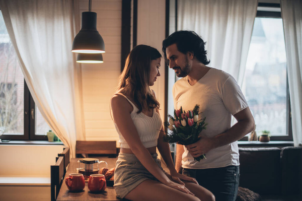 Couple aimant le soir dans le salon à la maison. Journée internationale de la femme. Un homme donne à une femme un bouquet de tulipes le 8 mars. Soirée romantique en famille à la maison. - Photo, image