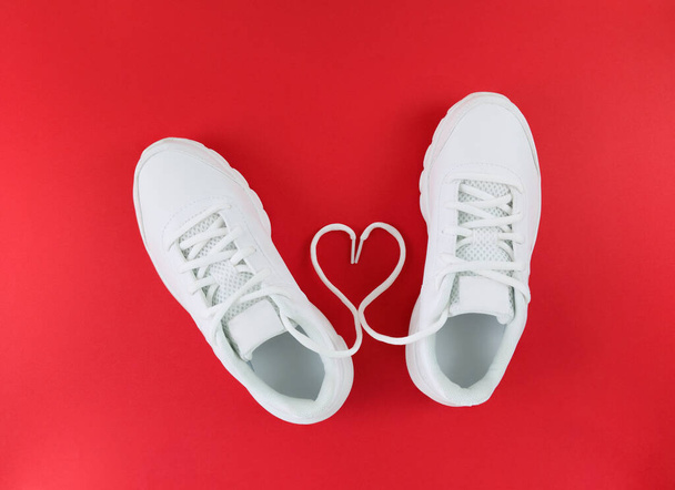 Weiße Sportschuhe und Herzform aus Schnürsenkeln auf rotem Hintergrund. Einfache flache Verlegung. - Foto, Bild
