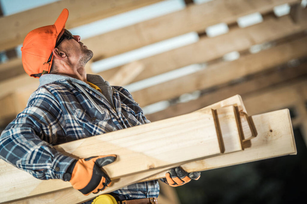 Construction Site Theme. Wooden House Structure Frame Work Job. Industrial Worker Moving Wooden Boards and Watching Up on the Roof Framing Structure. - Photo, Image