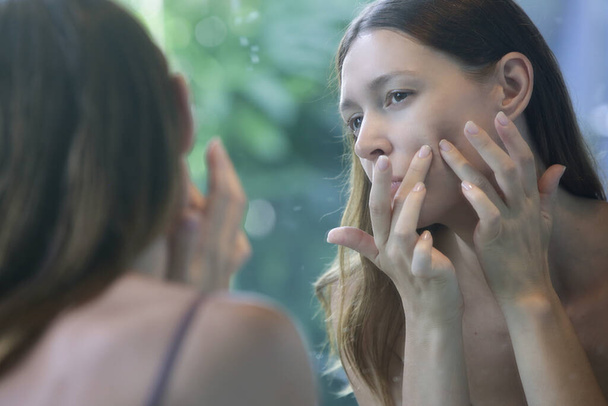 Portrait d'une belle jeune femme debout dans la salle de bain et examinant son visage dans le miroir, concept problématique de peau sujette à l'acné - Photo, image