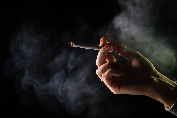 Woman hand holding cigarette in a smoke against black background. Smoking cannabis joint. Medical use. - Zdjęcie, obraz