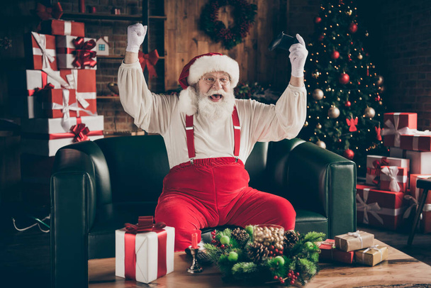 Photo of crazy addicted grey beard santa claus sit ouch have funny x-mas noel party play video game win raise fists wear red cap headwear in house indoors with christmas evergreen tree decoration - 写真・画像