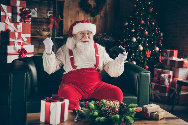 Excited addicted grey beard santa claus sit couch enjoy x-mas christmas event play video game win success raise fists wear cap headwear in house indoors noel evergreen tree ornament - Φωτογραφία, εικόνα
