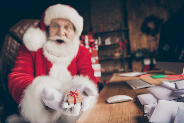 Blurred photo of shocked santa claus sit table showing hand small gift box demonstrate x-mas christmas present delivery wear red cap headwear in house indoors with noel decoration - Foto, afbeelding